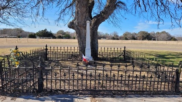 The Battle Creek burial ground near Dawson: a white marble shaft between two oaks, inside an iron fence, with a Texas flag at the base
