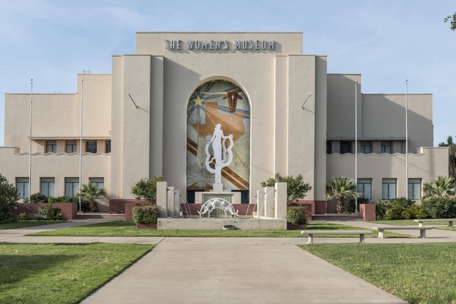 The front of the old Hall of Administration at Fair Park, later the Women's Museum: a cream art deco building with a tall arched mural and the white statue before it, a fountain and lawn in front