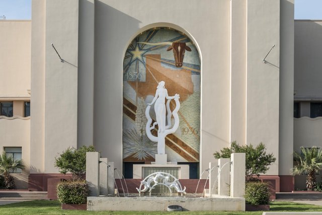 A white, stylised statue of a young woman rising out of a cactus, holding a sprig of cotton, in front of a tall arched mural of Texas with a lone star, a longhorn's head and the dates 1836 and 1936