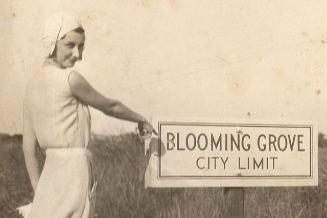 An old sepia photograph of a young woman in a white knotted cap and a long sleeveless white dress, looking back over her shoulder and pointing at a sign reading Blooming Grove City Limit beside tall grass