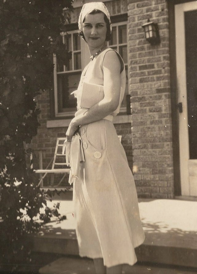 An old sepia photograph of the same young woman in a white knotted cap, beaded necklace and long white dress with large buttons, holding gloves, standing beside a brick house with a lantern by the door