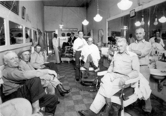 Inside a Blooming Grove barber shop: men in the barber chairs and on a bench in the foreground, and at the back, beside the raised shoe-shine stand, a tall thin Black man in a light jacket, Kildee