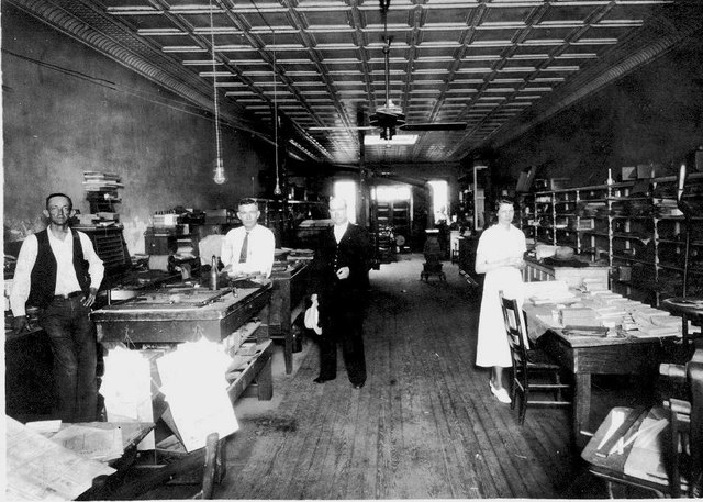 Four people in the newspaper office: a composing table at left, long tables stacked with printed sheets at right