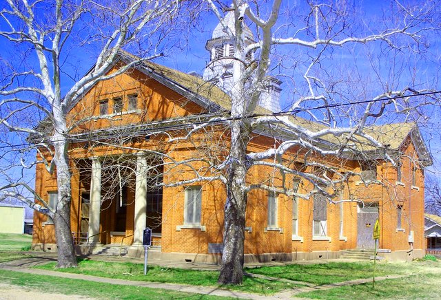 A two-story tan brick building with a columned portico and a white cupola, behind a pale sycamore