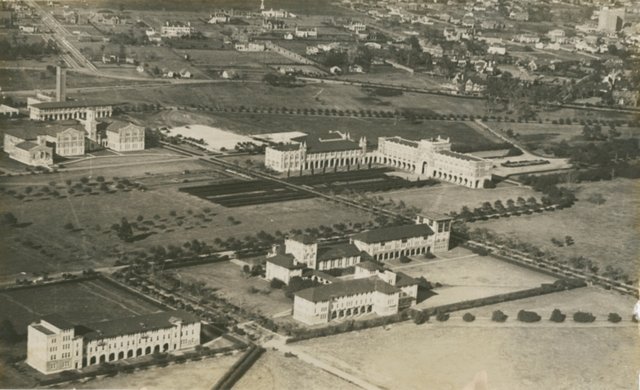 Aerial view of the Rice Institute campus in 1927: long arcaded buildings around a quadrangle, a tall tower at left, open fields and Houston's houses beyond