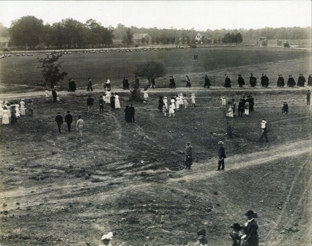 Faculty in caps and gowns walking in a long line across bare open ground, onlookers in white dresses and straw hats, the entrance gates in the distance