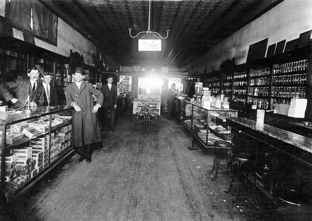 A drug store interior with glass cases, a pressed-tin ceiling and men standing at the counters