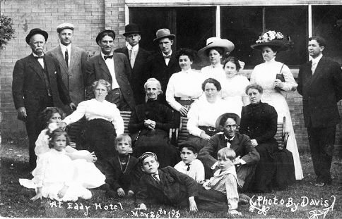 A group of men, women and children in their best clothes posed on a porch, the photograph inscribed At Eddy Hotel, May 1901, Photo by Davis