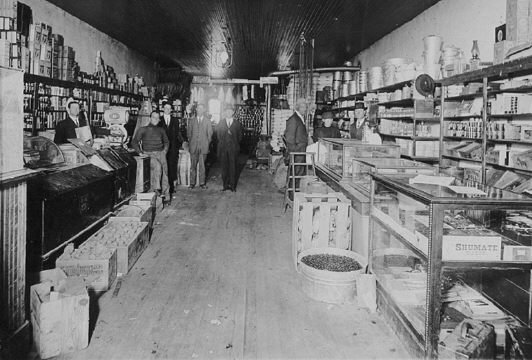 The long interior of a general store, shelves of goods to the ceiling, clerks and customers down the aisle