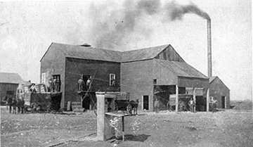 A cotton gin with a tall smokestack, mule wagons loaded with cotton waiting alongside