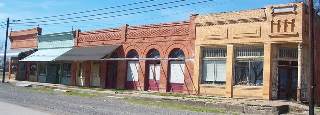 A row of old brick storefronts in Rice: two with metal awnings, a red brick front with four arched doorways, and a tan brick corner building with tall windows